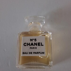 Puede incluir: Un peque&ntilde;o frasco de perfume rectangular con un tap&oacute;n transparente. El frasco contiene un l&iacute;quido &aacute;mbar claro y presenta una etiqueta blanca con texto negro: "N&deg;5 CHANEL PARIS EAU DE PARFUM."