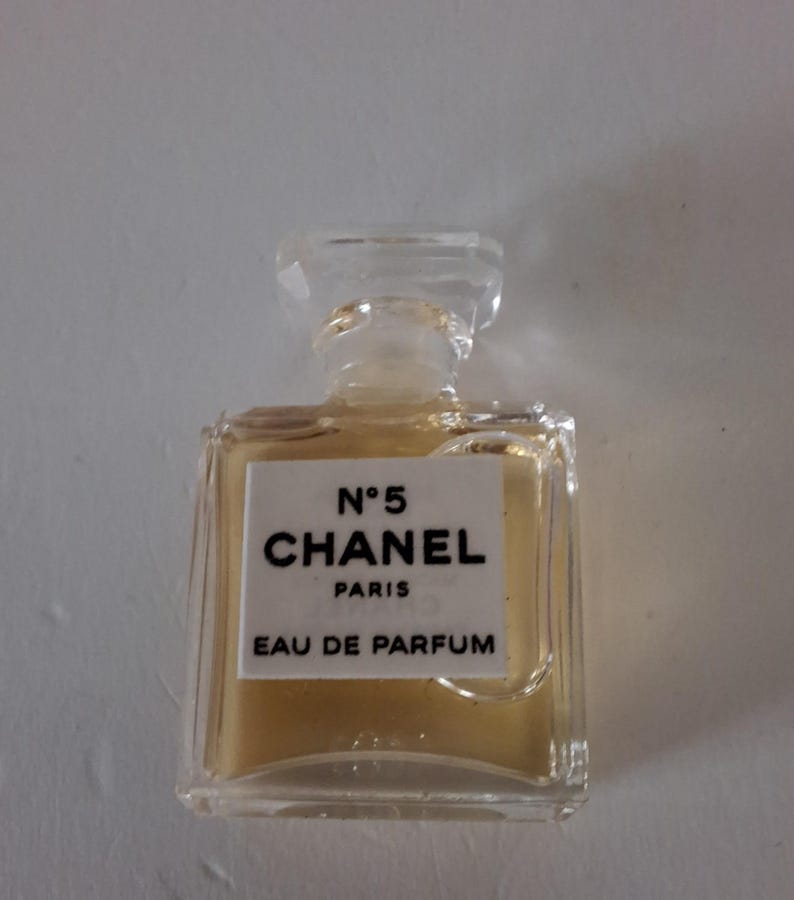 Puede incluir: Un peque&ntilde;o frasco de perfume rectangular con un tap&oacute;n transparente. El frasco contiene un l&iacute;quido &aacute;mbar claro y presenta una etiqueta blanca con texto negro: "N&deg;5 CHANEL PARIS EAU DE PARFUM."