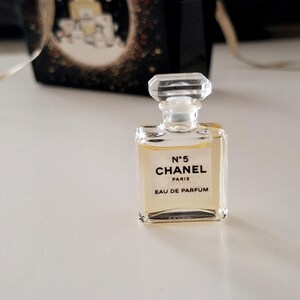 Puede incluir: Un peque&ntilde;o frasco de perfume de vidrio transparente, de forma rectangular y con tap&oacute;n facetado. Lleva la inscripci&oacute;n "N&deg;5 CHANEL PARIS EAU DE PARFUM". El l&iacute;quido interior es de color &aacute;mbar claro. En el fondo, una caja negra con detalles dorados.