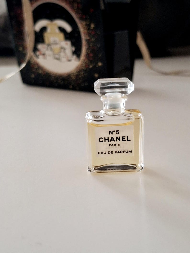 Puede incluir: Un peque&ntilde;o frasco de perfume de vidrio transparente, de forma rectangular y con tap&oacute;n facetado. Lleva la inscripci&oacute;n "N&deg;5 CHANEL PARIS EAU DE PARFUM". El l&iacute;quido interior es de color &aacute;mbar claro. En el fondo, una caja negra con detalles dorados.