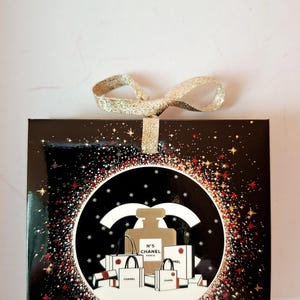 Puede incluir: Una caja de regalo negra con un lazo de cinta dorada. La caja presenta un dise&ntilde;o circular con un frasco de perfume, bolsas de compras y el texto "N&deg;5 CHANEL PARIS". El dise&ntilde;o est&aacute; rodeado de destellos de estrellas rojas y doradas.