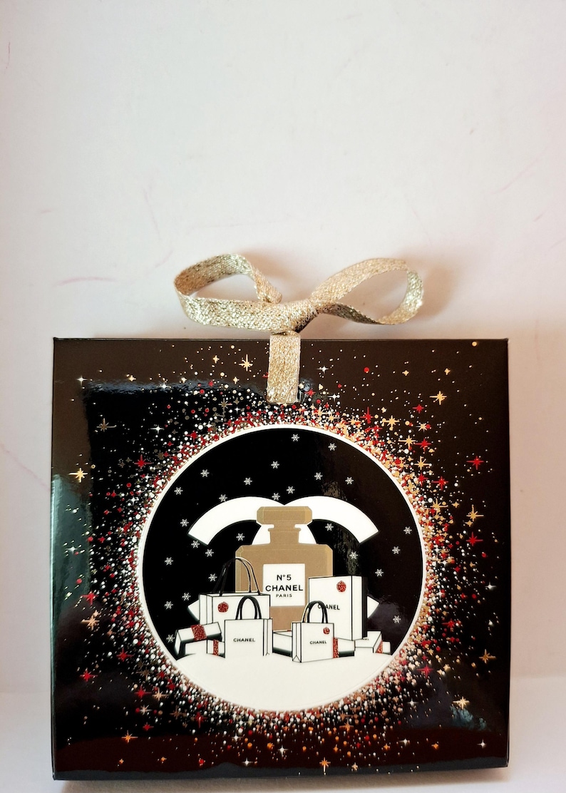 Puede incluir: Una caja de regalo negra con un lazo de cinta dorada. La caja presenta un dise&ntilde;o circular con un frasco de perfume, bolsas de compras y el texto "N&deg;5 CHANEL PARIS". El dise&ntilde;o est&aacute; rodeado de destellos de estrellas rojas y doradas.