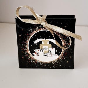 Puede incluir: Una caja de regalo negra con una cinta dorada atada con un lazo. La caja presenta un dise&ntilde;o circular con un frasco de perfume, bolsas de compras y el texto "N&deg;5 CHANEL PARIS" sobre un fondo de estrellas brillantes.