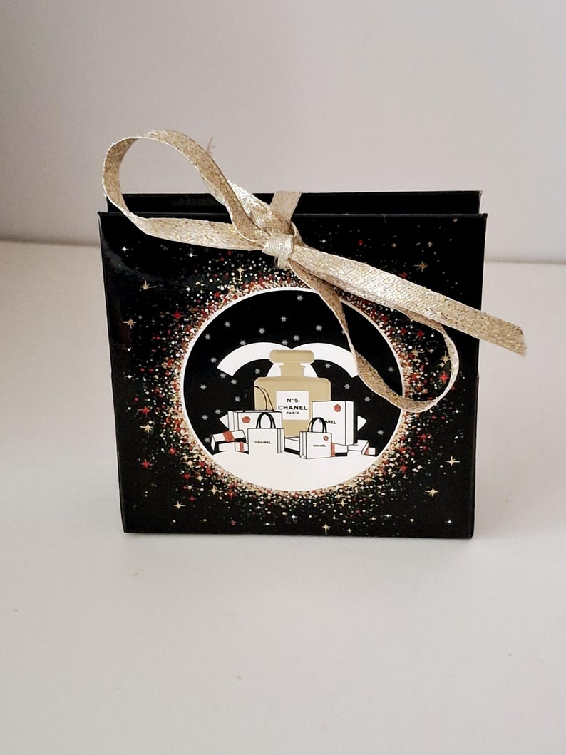 Puede incluir: Una caja de regalo negra con una cinta dorada atada con un lazo. La caja presenta un dise&ntilde;o circular con un frasco de perfume, bolsas de compras y el texto "N&deg;5 CHANEL PARIS" sobre un fondo de estrellas brillantes.