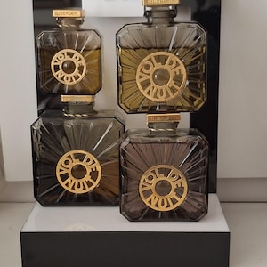 Könnte beinhalten: Vier quadratische Parfümflakons aus dunkelbraun getöntem Glas mit Strahlenmuster. Jeder Flakon hat ein kreisförmiges, goldfarbenes Metallemblem mit der Aufschrift Vol De Nuit und einen goldfarbenen Verschluss mit dem Markennamen Guerlain.