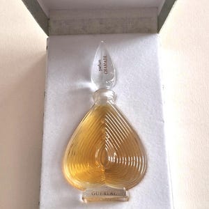Puede incluir: Un frasco de perfume de vidrio en forma de corazón con líquido ámbar, que presenta un tapón transparente con el texto 'parfum CHAMADE' y una base con la etiqueta 'GUERLAIN'. El frasco se presenta dentro de una caja rectangular abierta forrada en blanco.
