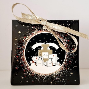 Puede incluir: Caja de regalo negra con lazo dorado. La caja presenta un dise&ntilde;o circular con un frasco de perfume, bolsas de compras y el logotipo de Chanel, sobre un fondo brillante.