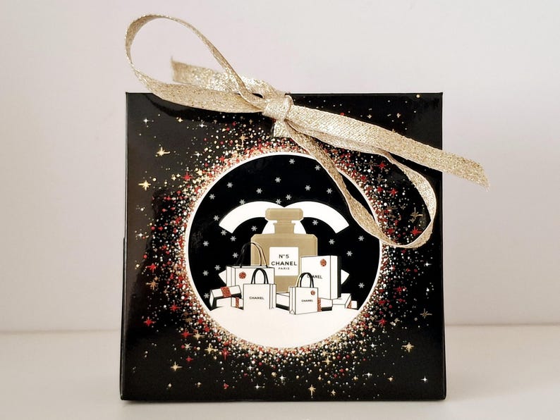 Puede incluir: Caja de regalo negra con lazo dorado. La caja presenta un dise&ntilde;o circular con un frasco de perfume, bolsas de compras y el logotipo de Chanel, sobre un fondo brillante.