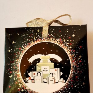 Puede incluir: Caja de regalo negra con lazo dorado brillante. La caja presenta un dise&ntilde;o circular con un frasco de perfume, bolsas de compras y el texto "N&deg;5 CHANEL PARIS". El dise&ntilde;o est&aacute; rodeado por un borde brillante.