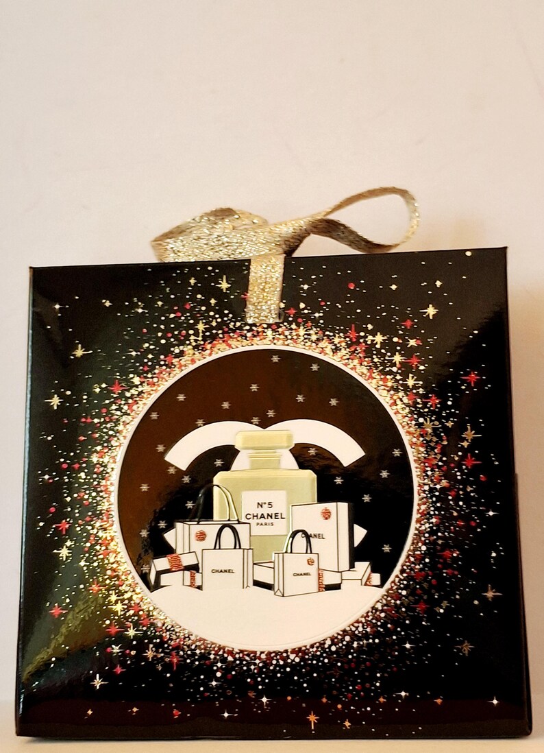 Puede incluir: Caja de regalo negra con lazo dorado brillante. La caja presenta un dise&ntilde;o circular con un frasco de perfume, bolsas de compras y el texto "N&deg;5 CHANEL PARIS". El dise&ntilde;o est&aacute; rodeado por un borde brillante.
