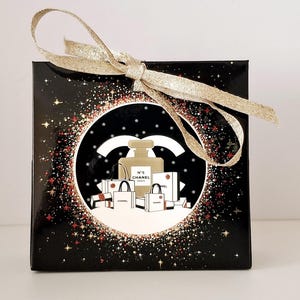 Puede incluir: Una caja de regalo negra con una cinta dorada atada con un lazo. La caja presenta un dise&ntilde;o circular con un frasco de perfume, bolsas de compras y el logotipo de Chanel. El dise&ntilde;o est&aacute; sobre un fondo negro con acentos de estrellas doradas y rojas.