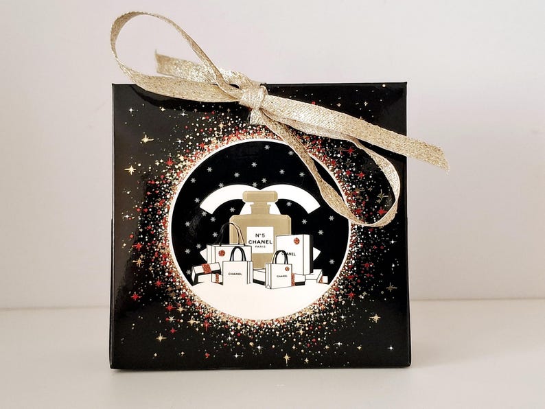 Puede incluir: Una caja de regalo negra con una cinta dorada atada con un lazo. La caja presenta un dise&ntilde;o circular con un frasco de perfume, bolsas de compras y el logotipo de Chanel. El dise&ntilde;o est&aacute; sobre un fondo negro con acentos de estrellas doradas y rojas.