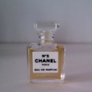 Puede incluir: Un peque&ntilde;o frasco de perfume rectangular con un tap&oacute;n transparente cuadrado. El frasco contiene un l&iacute;quido &aacute;mbar claro y presenta una etiqueta blanca con el texto "N&deg;5 CHANEL PARIS EAU DE PARFUM" en letras negras. El fondo est&aacute; borroso.