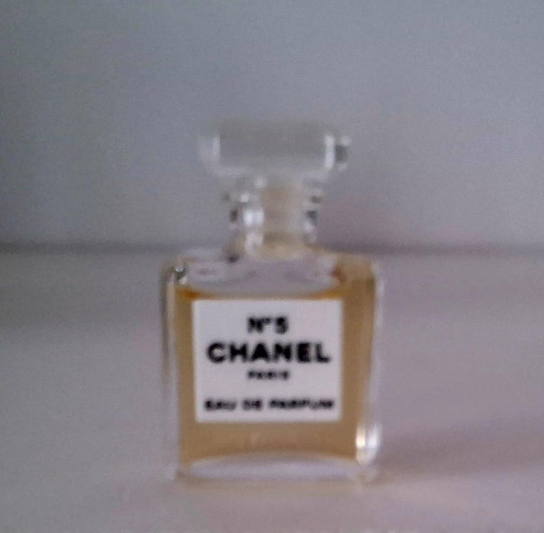 Puede incluir: Un peque&ntilde;o frasco de perfume rectangular con un tap&oacute;n transparente cuadrado. El frasco contiene un l&iacute;quido &aacute;mbar claro y presenta una etiqueta blanca con el texto "N&deg;5 CHANEL PARIS EAU DE PARFUM" en letras negras. El fondo est&aacute; borroso.