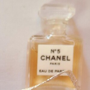 Puede incluir: Un peque&ntilde;o frasco de perfume rectangular con un tap&oacute;n transparente facetado. El frasco contiene un l&iacute;quido &aacute;mbar claro y presenta una etiqueta blanca con el texto "N&deg;5 CHANEL PARIS EAU DE PARFUM". Una cinta plateada est&aacute; en primer plano.