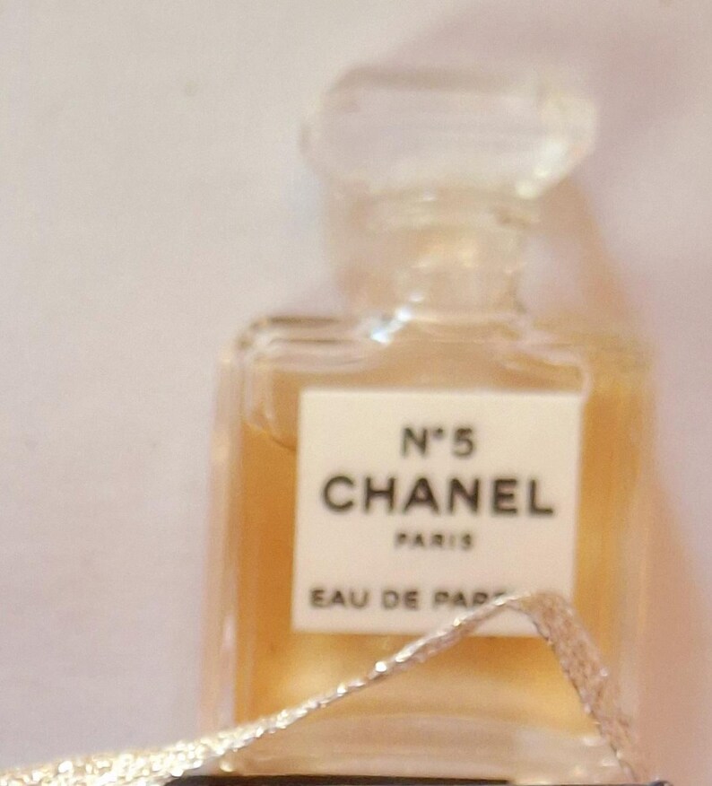 Puede incluir: Un peque&ntilde;o frasco de perfume rectangular con un tap&oacute;n transparente facetado. El frasco contiene un l&iacute;quido &aacute;mbar claro y presenta una etiqueta blanca con el texto "N&deg;5 CHANEL PARIS EAU DE PARFUM". Una cinta plateada est&aacute; en primer plano.