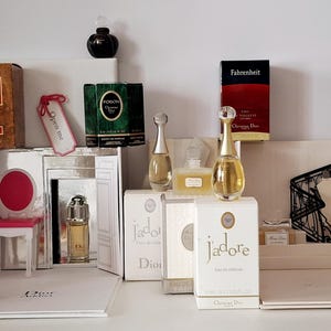 Peut inclure: Une collection de flacons et boîtes de parfum Christian Dior, incluant Eau Sauvage, Poison, Fahrenheit et J'adore. L'ensemble présente des flacons miniatures, des emballages de marque, une petite chaise rose et une illustration d'escalier.