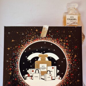 Puede incluir: Una caja de regalo negra con una cinta dorada brillante y un dise&ntilde;o circular con un frasco de perfume y bolsas de compras. El dise&ntilde;o est&aacute; rodeado de purpurina roja y dorada. Un frasco de perfume es visible en el fondo.