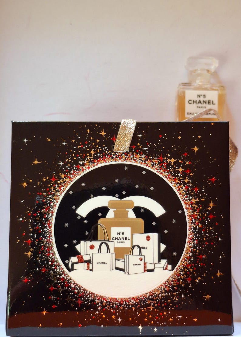 Puede incluir: Una caja de regalo negra con una cinta dorada brillante y un dise&ntilde;o circular con un frasco de perfume y bolsas de compras. El dise&ntilde;o est&aacute; rodeado de purpurina roja y dorada. Un frasco de perfume es visible en el fondo.