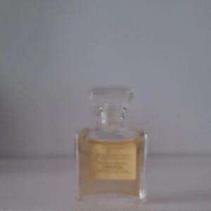 Puede incluir: Un peque&ntilde;o frasco de perfume transparente con forma rectangular y tap&oacute;n transparente. El frasco contiene un l&iacute;quido de color &aacute;mbar claro y tiene una etiqueta dorada. El frasco est&aacute; sobre una superficie blanca.