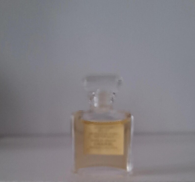 Puede incluir: Un peque&ntilde;o frasco de perfume transparente con forma rectangular y tap&oacute;n transparente. El frasco contiene un l&iacute;quido de color &aacute;mbar claro y tiene una etiqueta dorada. El frasco est&aacute; sobre una superficie blanca.