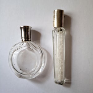 Puede incluir: Dos frascos de perfume de vidrio vintage. Uno es redondo con una tapa plateada, el otro es rectangular con una tapa plateada y lleno de cristales blancos. Ambos frascos están vacíos.