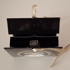 Puede incluir: Una caja de anillos negra con una cinta dorada y un anillo con una gema grande y transparente. El anillo se muestra dentro de la caja abierta, sobre un fondo blanco. La caja est&aacute; abierta, revelando el anillo.