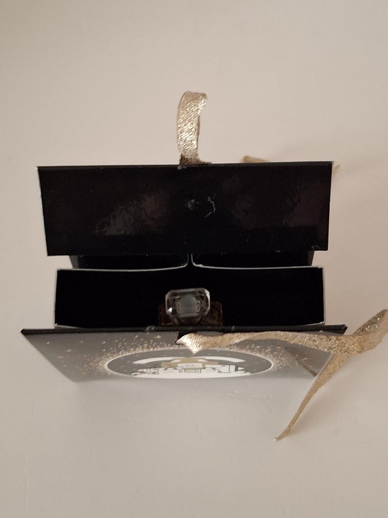 Puede incluir: Una caja de anillos negra con una cinta dorada y un anillo con una gema grande y transparente. El anillo se muestra dentro de la caja abierta, sobre un fondo blanco. La caja est&aacute; abierta, revelando el anillo.