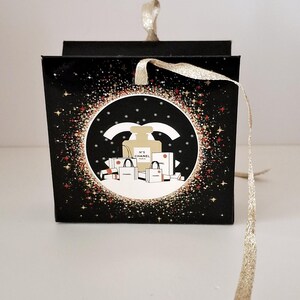 Puede incluir: Caja de regalo negra con una cinta dorada. La caja presenta un dise&ntilde;o circular con un frasco de perfume, bolsas de compras y el texto "N&deg;5 CHANEL PARIS". El dise&ntilde;o est&aacute; rodeado de purpurina dorada y roja.