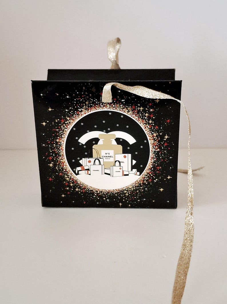 Puede incluir: Caja de regalo negra con una cinta dorada. La caja presenta un dise&ntilde;o circular con un frasco de perfume, bolsas de compras y el texto "N&deg;5 CHANEL PARIS". El dise&ntilde;o est&aacute; rodeado de purpurina dorada y roja.