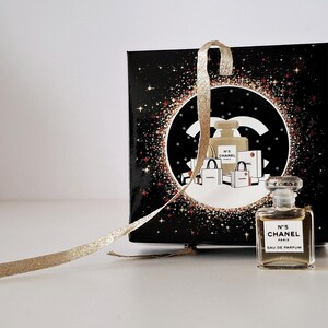 Puede incluir: Un peque&ntilde;o frasco de Chanel No. 5 Eau de Parfum junto a una caja de regalo negra con una cinta dorada. La caja presenta un dise&ntilde;o circular con el logo de Chanel e ilustraciones de frascos de perfume y bolsas de compras.