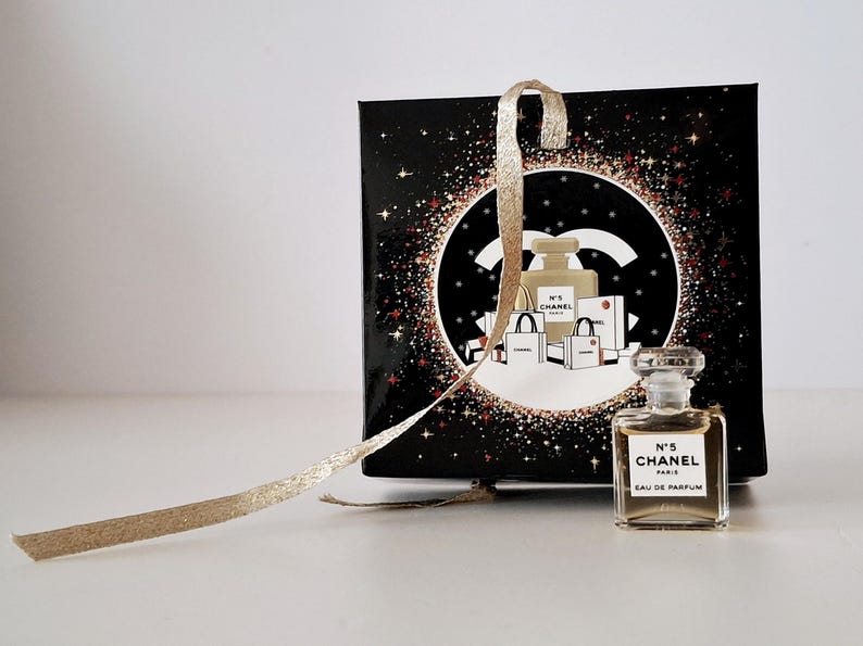 Puede incluir: Un peque&ntilde;o frasco de Chanel No. 5 Eau de Parfum junto a una caja de regalo negra con una cinta dorada. La caja presenta un dise&ntilde;o circular con el logo de Chanel e ilustraciones de frascos de perfume y bolsas de compras.