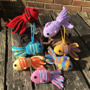 May include: Six crocheted fish toys in various colours, including red, purple, blue, yellow, orange, and pink. The fish are all different sizes and have different patterns. They are all hanging from a wooden branch.