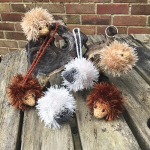May include: Five crocheted hedgehog ornaments in various colours, including brown, white, and grey. The hedgehogs are sitting on a wooden surface.
