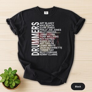 May include: Black t-shirt featuring the word "DRUMMERS" in white, alongside a list of names. A small drum set graphic is visible. The shirt is made of a soft material. The color is black.