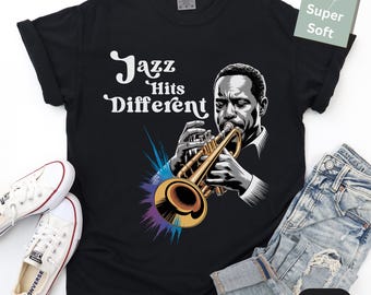 May include: Black t-shirt with the text "Jazz Hits Different" and an illustration of a musician playing a trumpet. The trumpet is gold-colored, and the illustration has a colorful burst design.