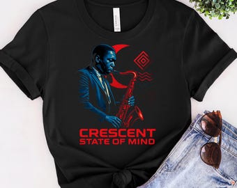 May include: Black t-shirt featuring a blue and red illustration of a musician playing a saxophone. The text "CRESCENT STATE OF MIND" is printed in red below the illustration. The shirt is made of a soft material.