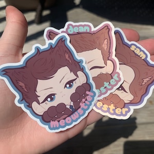 Puede incluir: Tres pegatinas coloridas con personajes de dibujos animados con orejas de gato. Las pegatinas tienen los nombres "Dean", "Sam" y "Castiel" escritos en ellas.