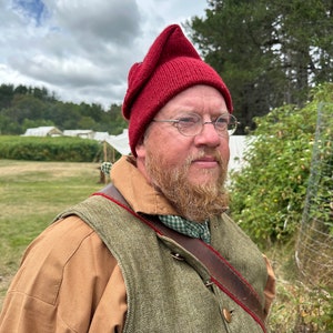 May include: A person wearing a burgundy knitted cap with a pointed top, round spectacles, and a beard. They are wearing a brown shirt, a green waistcoat with red trim, and a leather strap. The background shows tents and trees.
