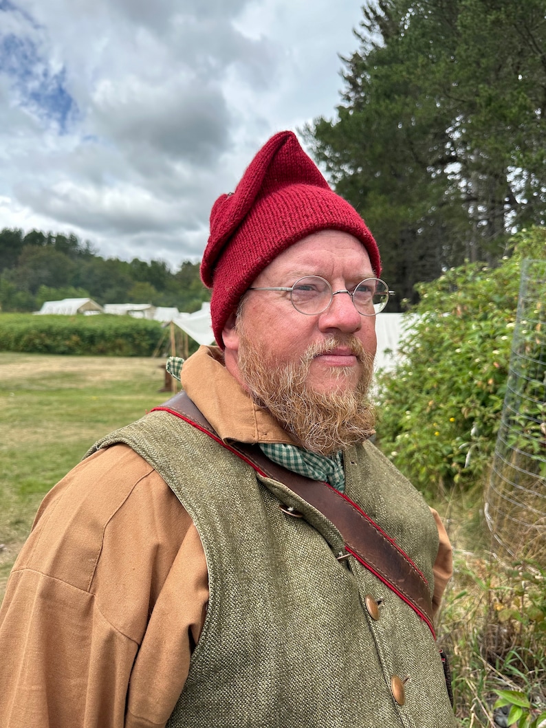 May include: A person wearing a burgundy knitted cap with a pointed top, round spectacles, and a beard. They are wearing a brown shirt, a green waistcoat with red trim, and a leather strap. The background shows tents and trees.