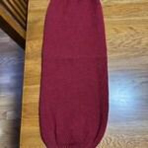 May include: A burgundy knitted tube, possibly a leg warmer or sleeve, rests on a wooden surface. The item is made from thick, textured yarn, and the colour is a deep, solid hue.