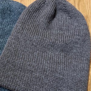 May include: Close-up of two knitted beanies. One is a dark grey colour, and the other is a muted blue. The beanies are made of a ribbed knit fabric. The background is a light brown wooden surface.