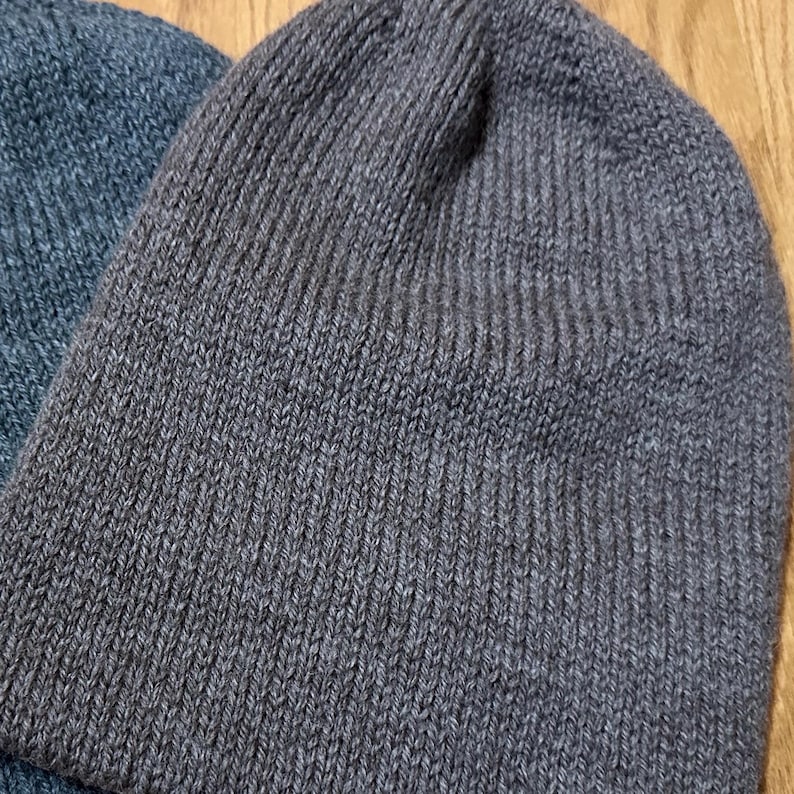 May include: Close-up of two knitted beanies. One is a dark grey colour, and the other is a muted blue. The beanies are made of a ribbed knit fabric. The background is a light brown wooden surface.