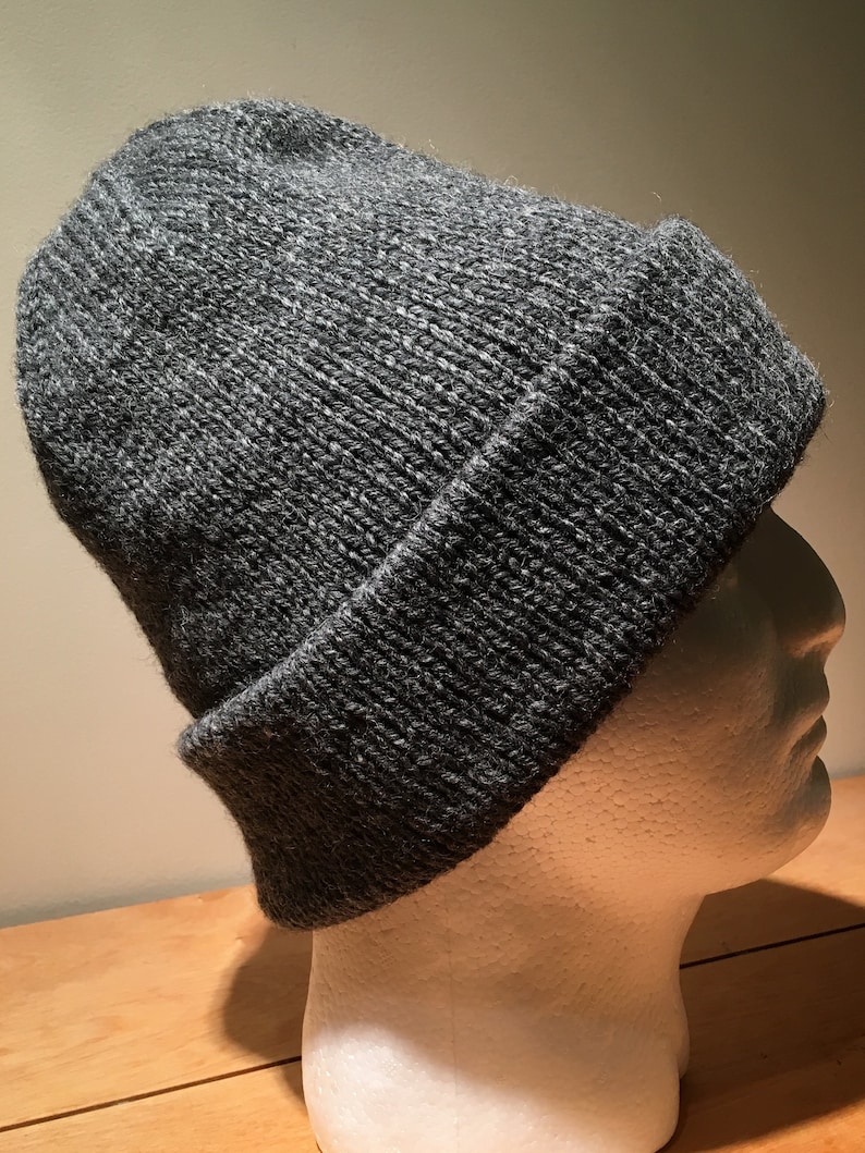 May include: A dark grey knitted beanie hat with a folded-over brim. The hat is made from textured yarn and is displayed on a white mannequin head. A classic winter accessory.
