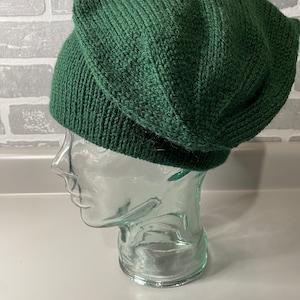 May include: A dark green knitted beanie hat. The hat is made of a thick, textured yarn and has a folded brim. The hat is displayed on a clear glass head form against a white and grey background.