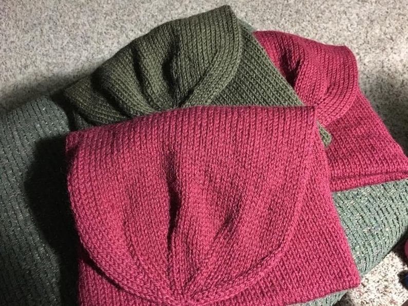 May include: Close-up of three knitted items in shades of green and red. The items appear to be folded, with a flap-like design. The texture of the knitwear is visible, with vertical ribbing. The items are likely accessories or small garments.