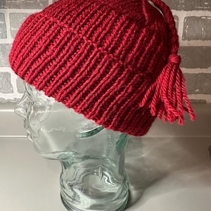May include: A vibrant red knit beanie with a ribbed texture. The hat features a tassel detail on the side, adding a playful touch. The beanie is displayed on a clear glass head form.