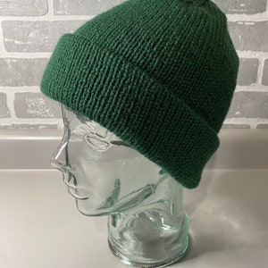 May include: A dark green knitted beanie hat with a turned-up brim. The hat is displayed on a clear glass head form against a grey brick wall background. The beanie is made of a ribbed knit fabric.
