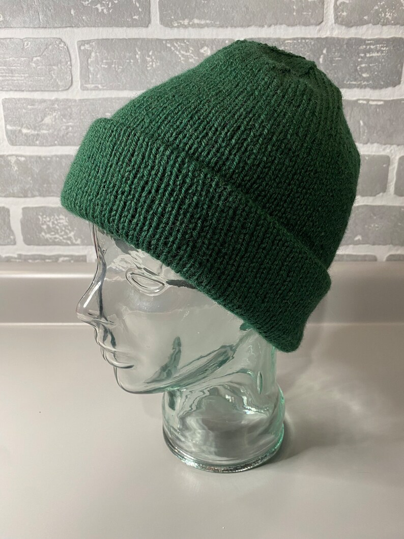 May include: A dark green knitted beanie hat with a turned-up brim. The hat is displayed on a clear glass head form against a grey brick wall background. The beanie is made of a ribbed knit fabric.
