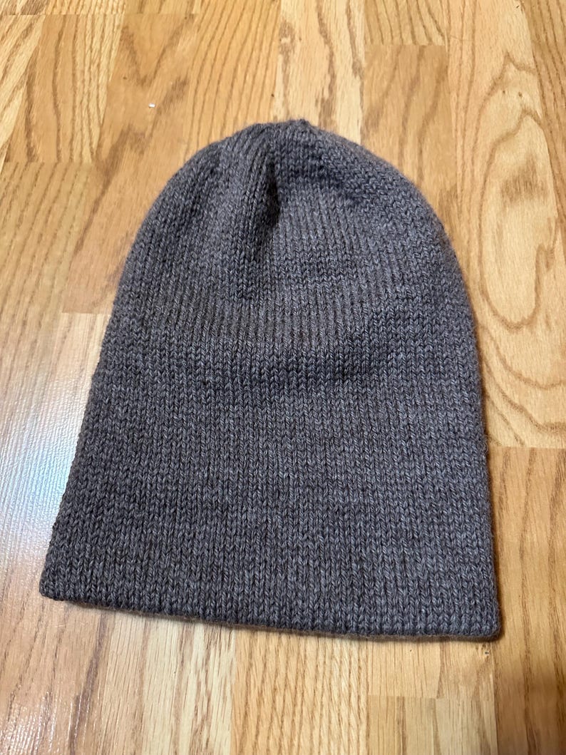 May include: A grey knitted beanie hat. The hat is a solid colour and has a ribbed texture. The beanie is a simple, classic design, suitable for cold weather. The hat is lying on a wooden surface.